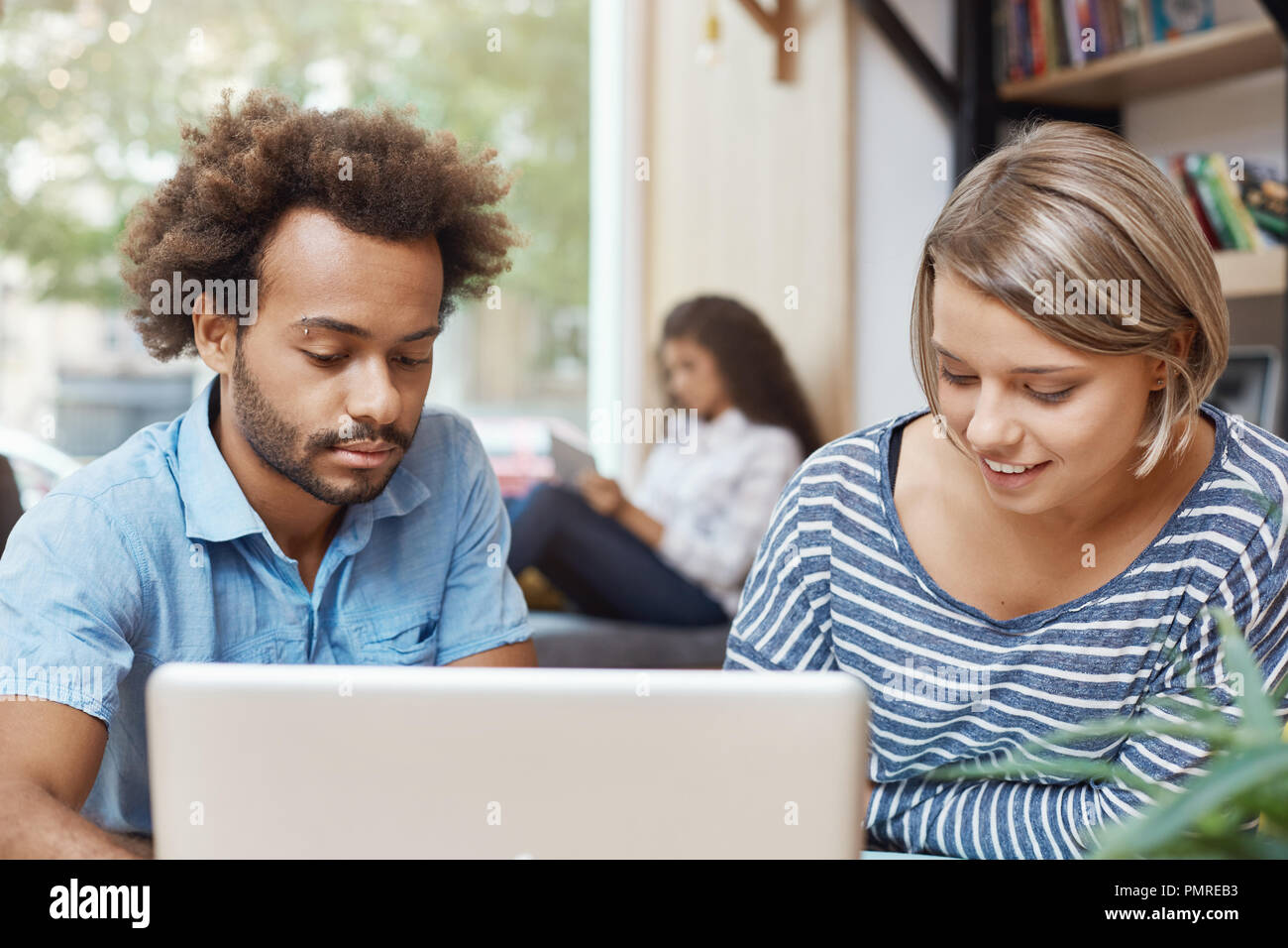 Two young freelance designers sitting in co-working space, looking in laptop monitor, searching for new ideas for furniture designers. Pair of young cheerful people smiling, spending, morning in library. Stock Photo