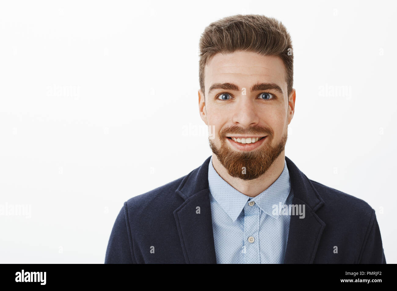Ambitious handsome and young stylish mature male with beard and big blue eyes smiling excited and pleased grin standing in trendy suit over gray wall waiting for chance show skills over gray wall Stock Photo