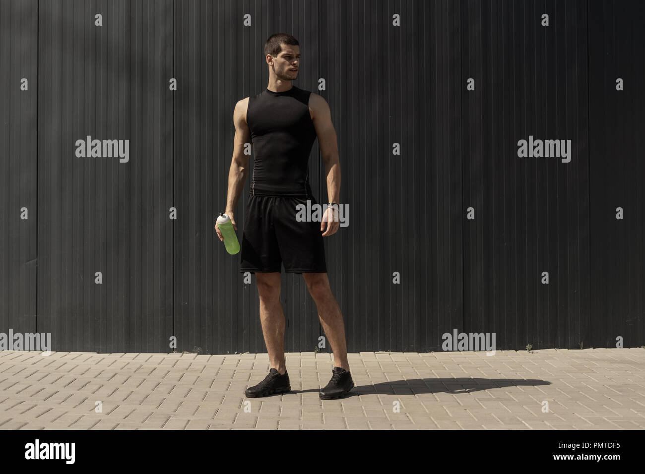 Athletic modern young man posing on a gray metallic wall with a water bottle in his hand. Hydration and nutrition's concept. Fitness muscled male. Hum Stock Photo