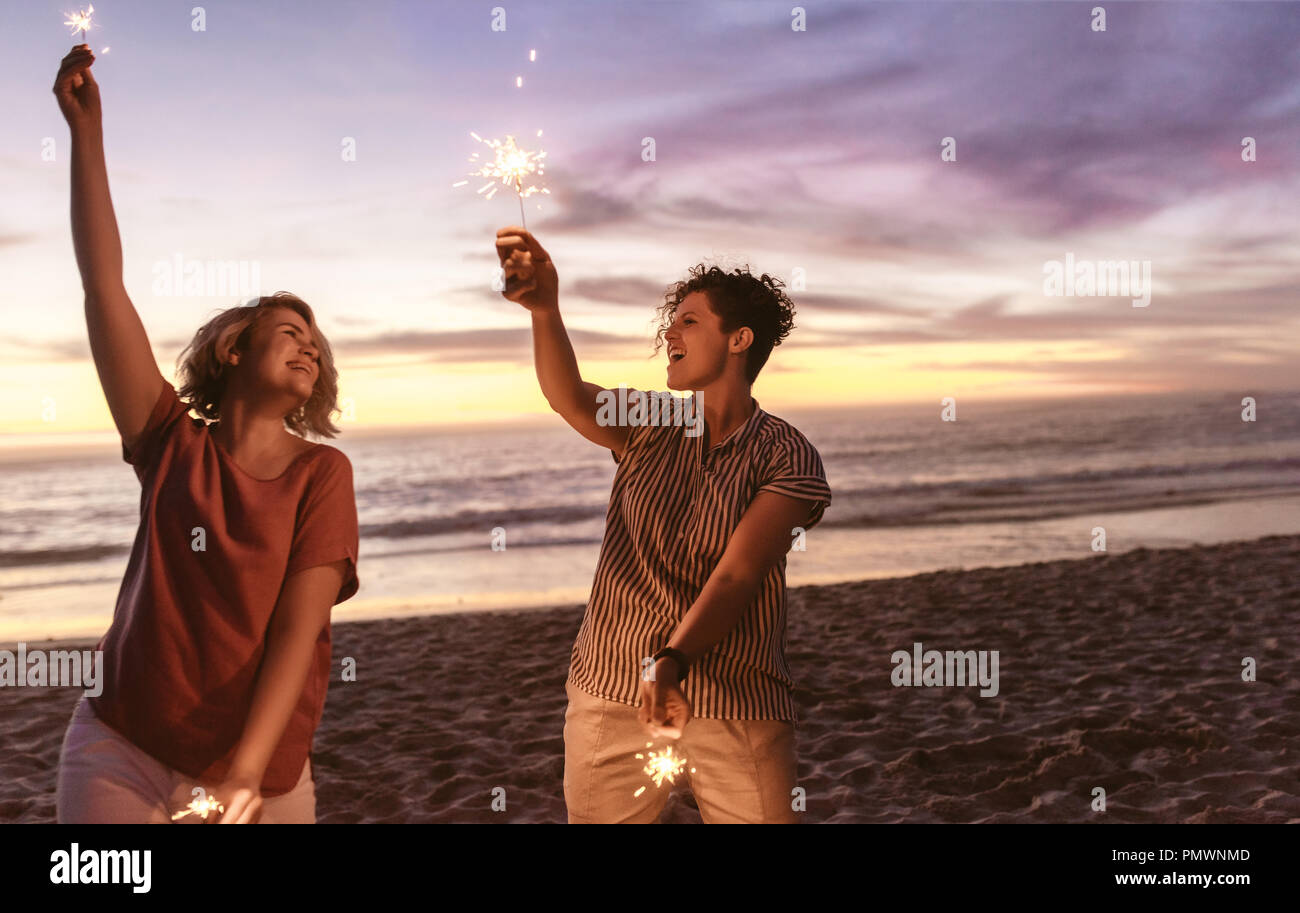 Two laughing young female friends standing side by side together on a beach at dusk having fun with sparklers Stock Photo