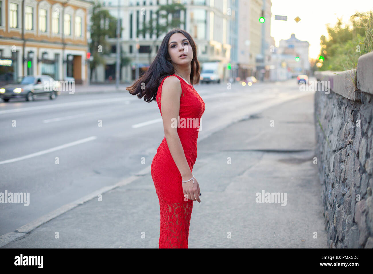 Beautiful girl in a red dress is standing near the roadway of the city ...