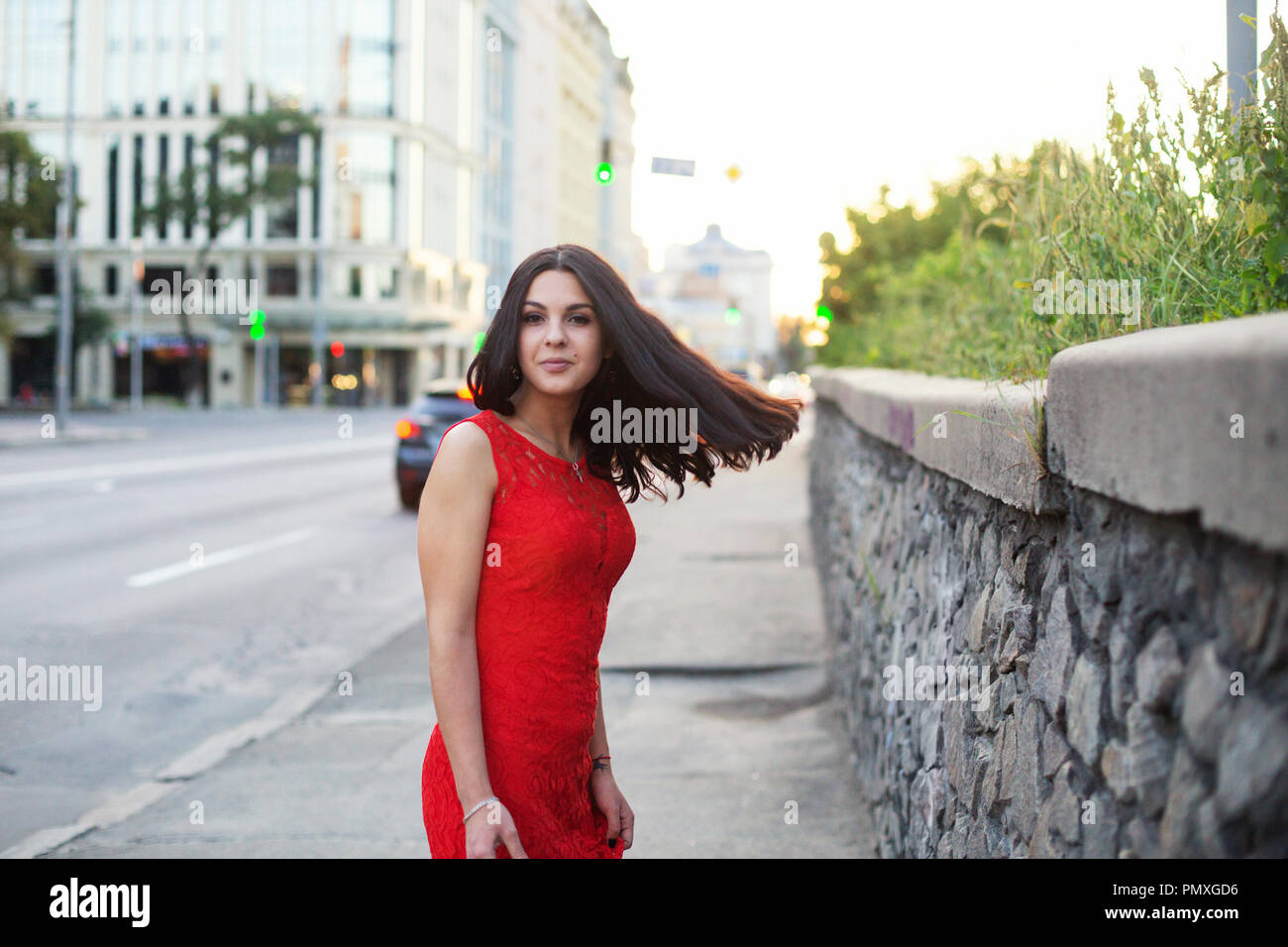 Beautiful girl in a red dress is standing near the roadway of the city ...