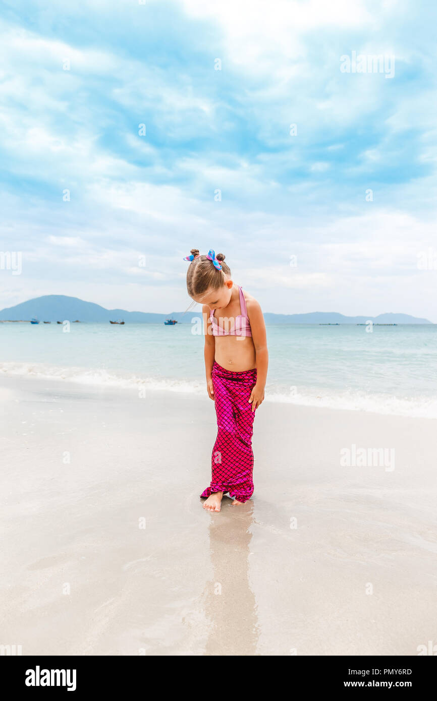 Little beautiful girl dressed in swimsuit as a mermaid stands on the seashore. Stock Photo