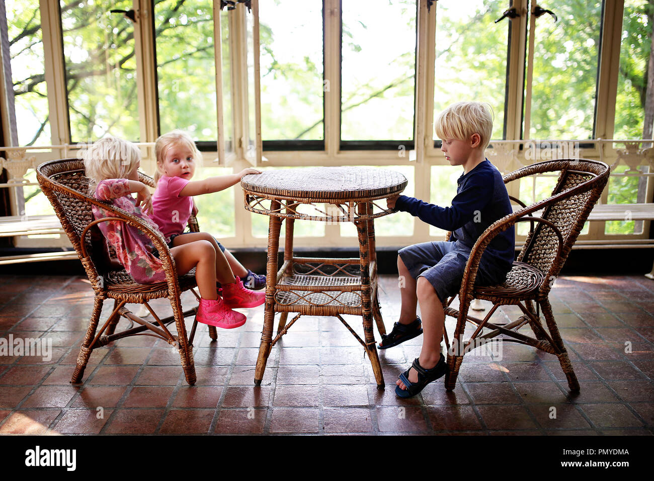 Three little children are sitting at an old wicker bistro table in a sun room patio, waiting for their food to be served. Stock Photo