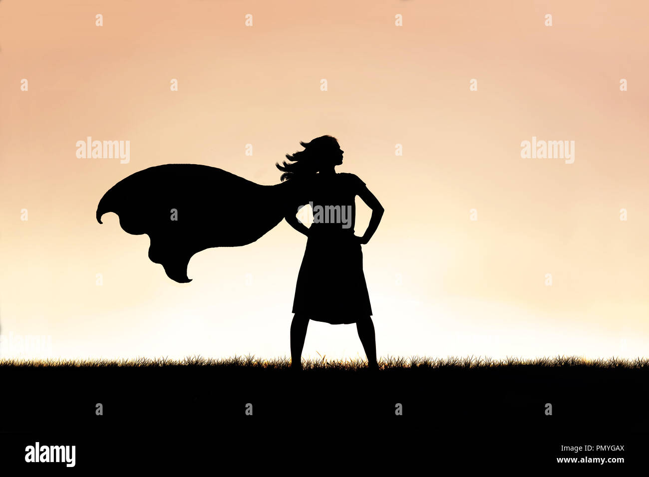 The silhouette of a strong, beautiful caped super hero woman stands isolated against a sunset in the sky background. Stock Photo