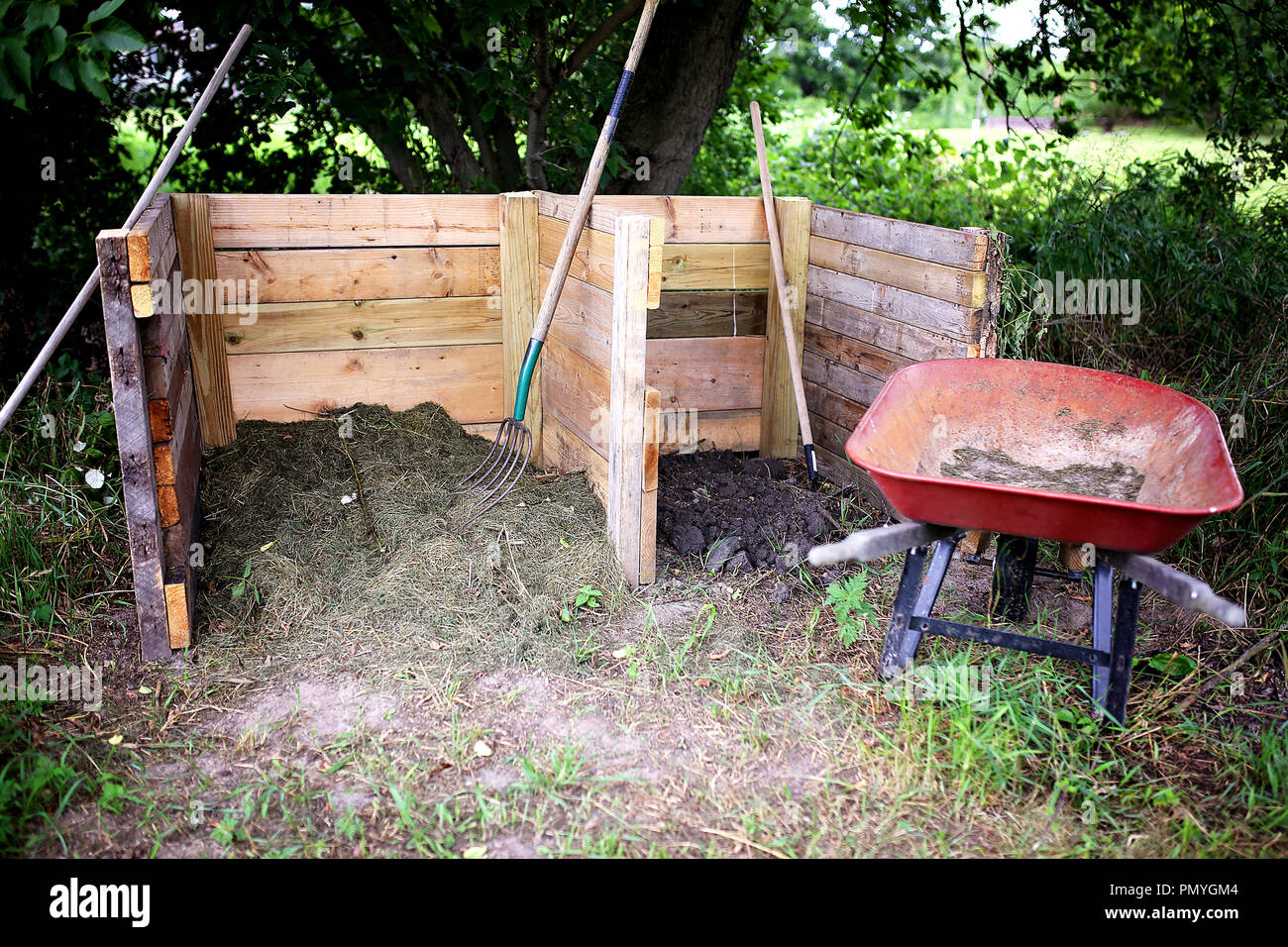 A homemade wooden 2 stage compost bin is constructed in a home garden, with a wheelbarrow and pitchfork sitting close by. Stock Photo