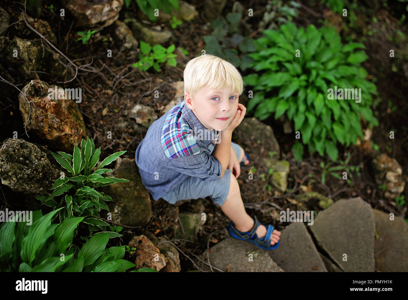A peaceful young, 6 year old boy is sitting in a rock garden by some green hosta plants on a sumemr day. Stock Photo