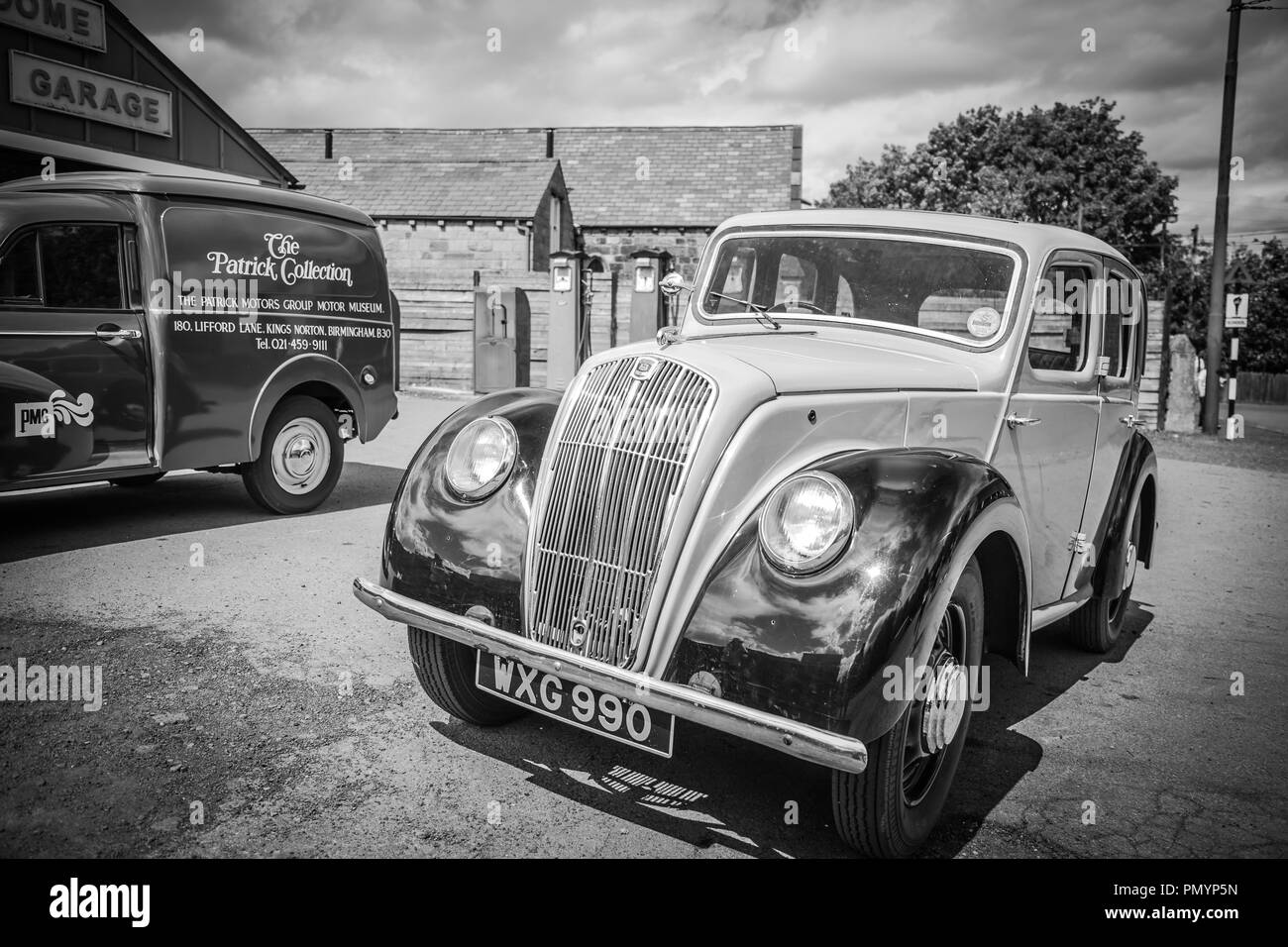 Monochrome view of vintage vehicles parked outside vintage UK garage. Vintage petrol pumps in background. Stock Photo