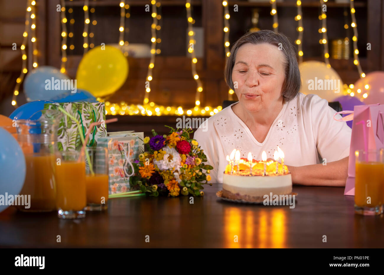 Elderly woman celebrating her anniversary at home. Senior woman blowing candles on birthday cake. Celebrating aging concept. Stock Photo