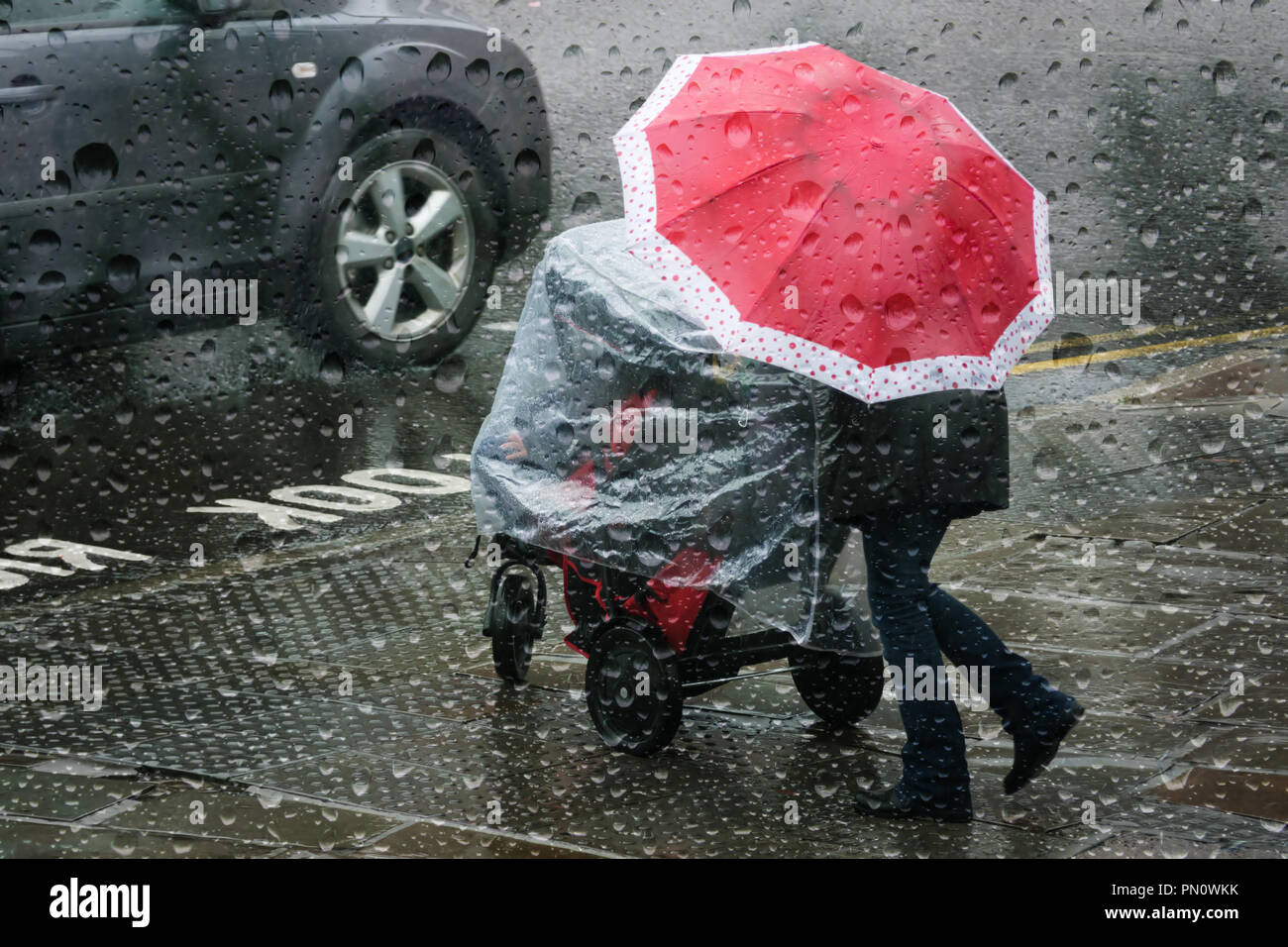 Looking through a raindrop covered window at a woman pushing a child's buggy getting caught in heavy autumn rain shower with an umbrella. Stock Photo