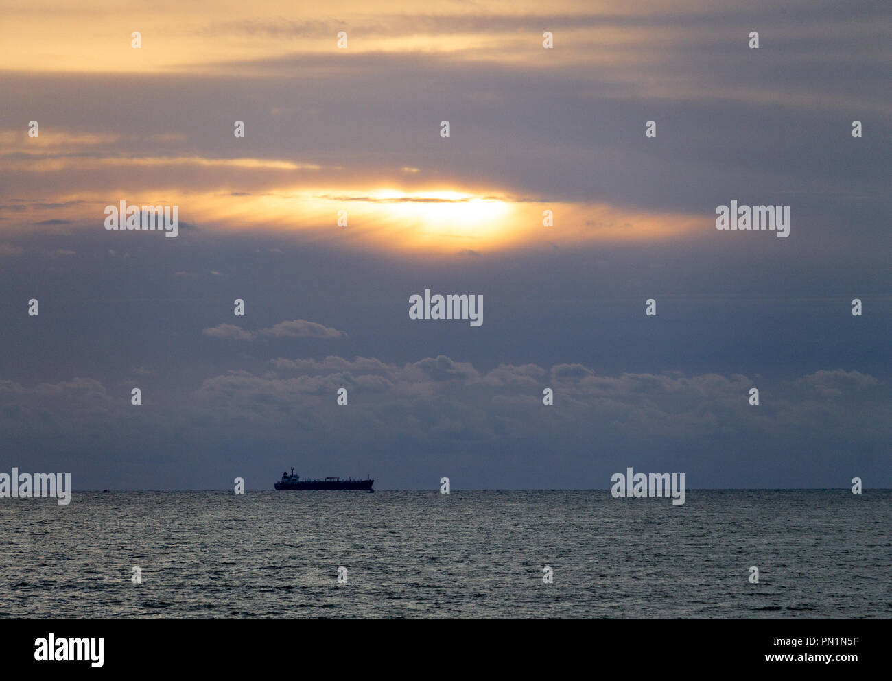 A distant cargo ship is seen on the ocean at the horizon, with the sun peaking through the clouds. Stock Photo