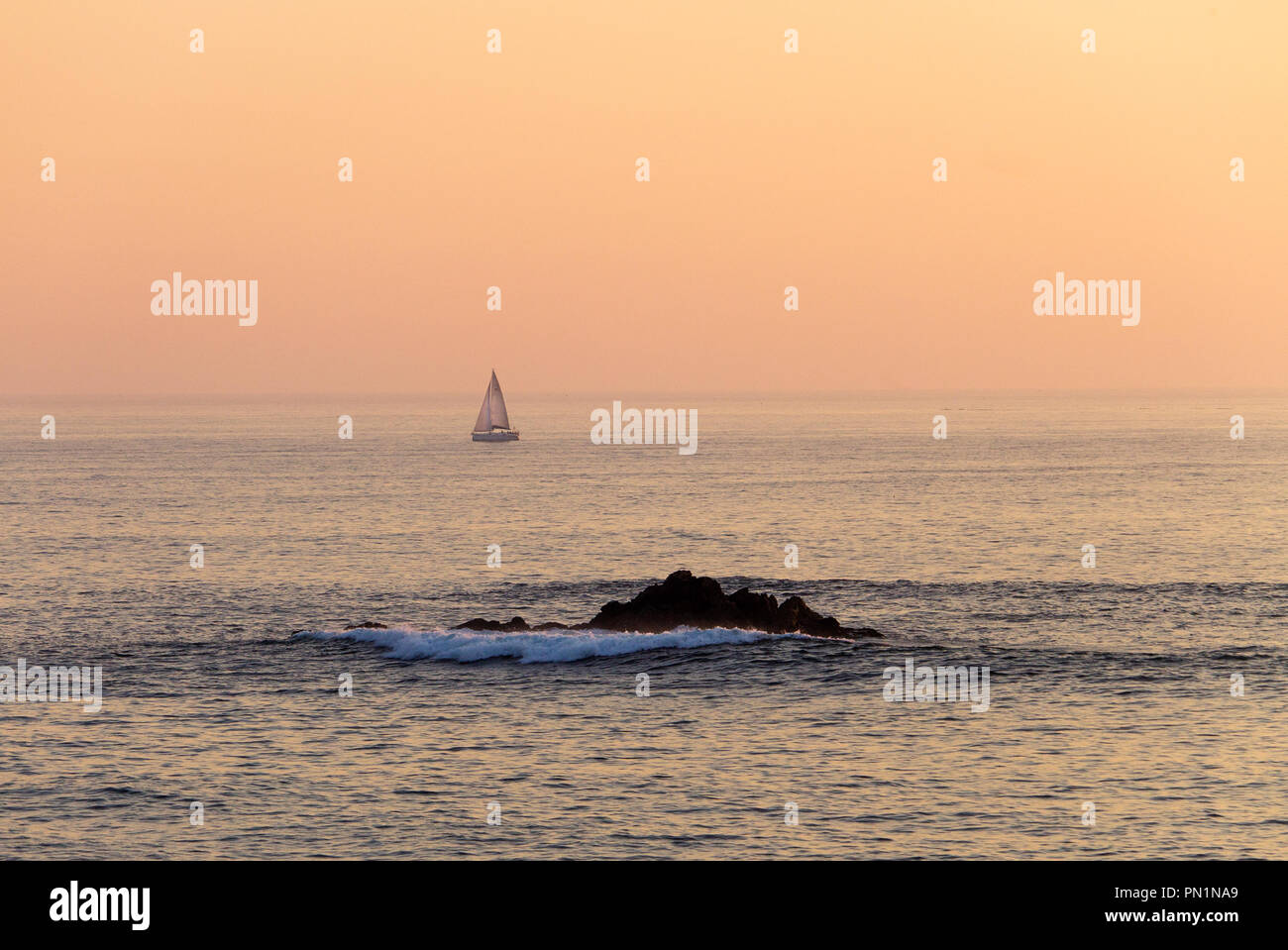 A distant sailboat sails along the sea during sunset with some waves crashing on a rock formation, with the orange colored sky in the background. Stock Photo