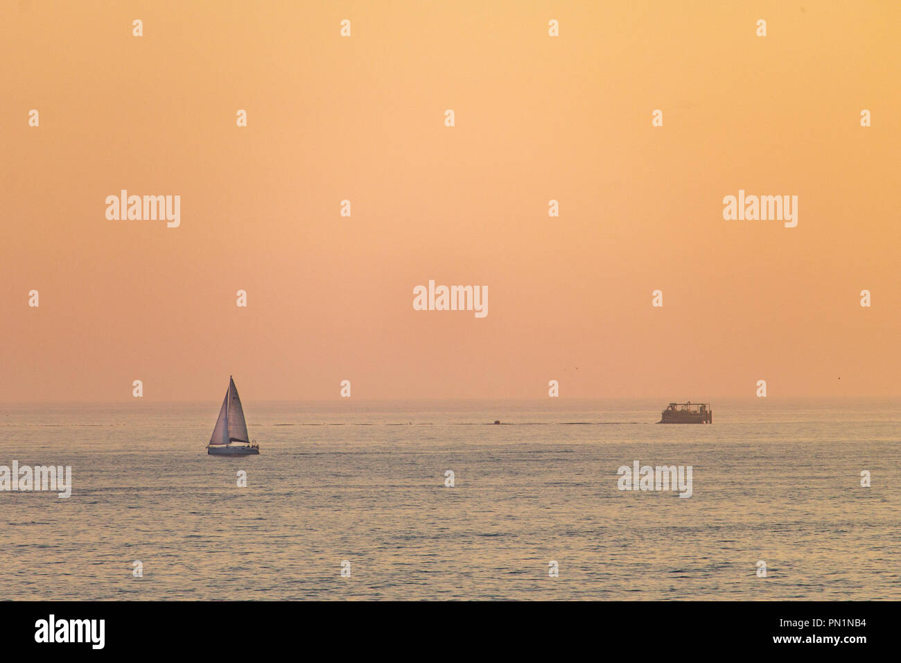 A distant sailboat sails along the sea during sunset, with the orange colored sky in the background. Stock Photo