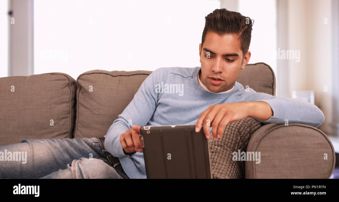 Young Hispanic man sitting on couch using tablet computer in living room Stock Photo