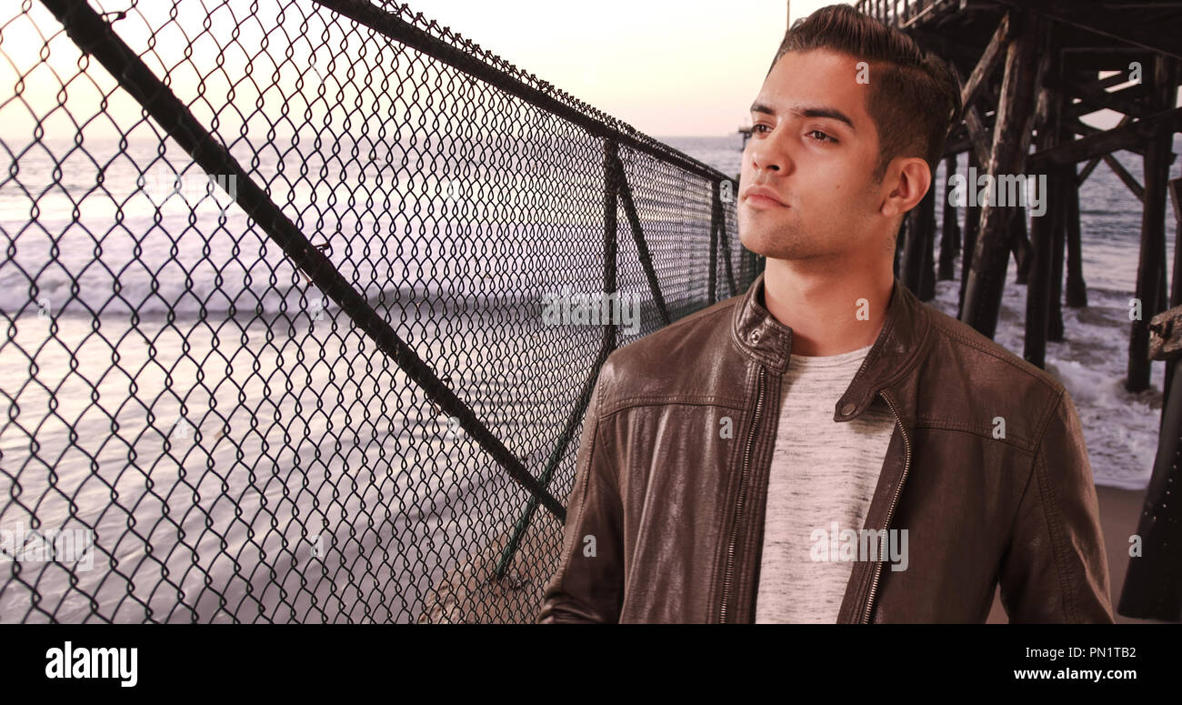 Fashionable millennial Hispanic man standing under pier on the beach Stock Photo