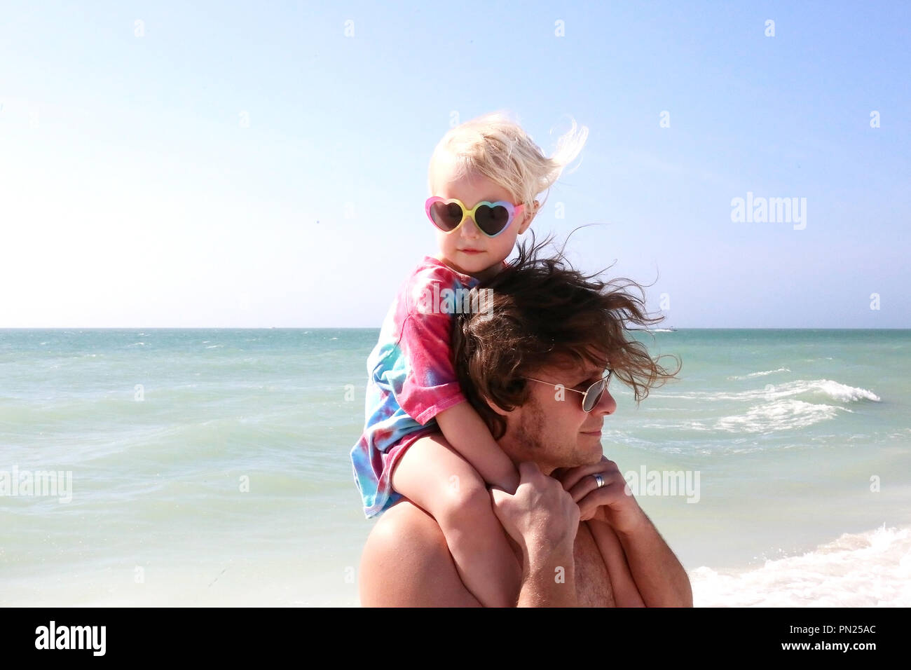 A Cute 2 Year Old Toddler Girl Is Sitting On Her Father S Shoulders Wearing Rainbow Heart Shaped Glasses On A Windy Day At The Beach On The Ocean Stock Photo Alamy alamy