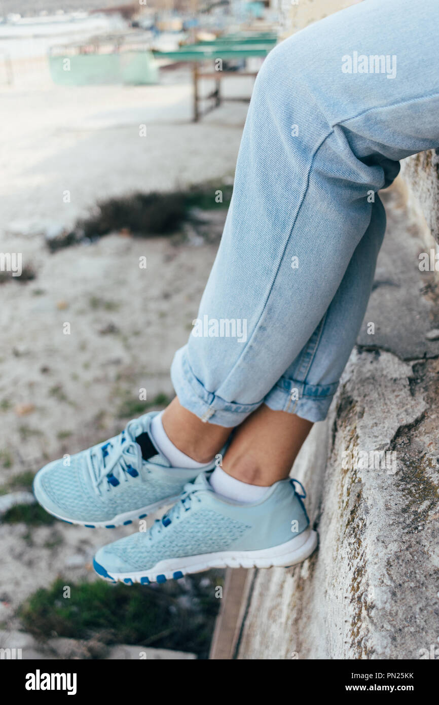 Close-up of female legs in blue rolled up jeans and sneakers. Young woman  sitting on high concrete parapet in the city, vertical framing Stock Photo  - Alamy