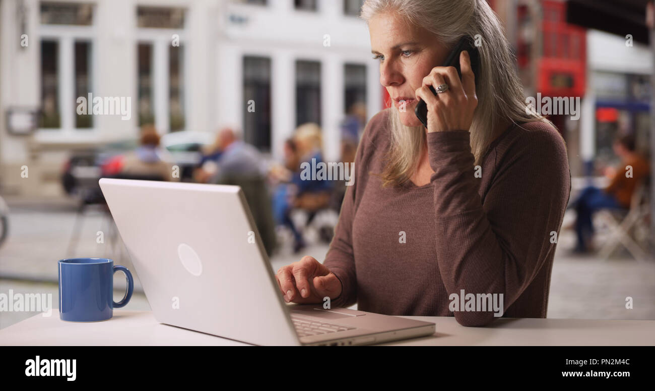 Busy older white woman works from laptop and makes phone call at cafe Stock Photo