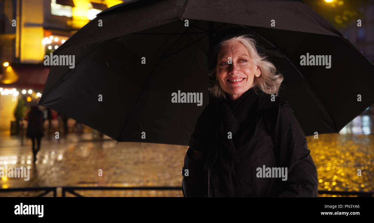 Senior woman in Paris standing under umbrella on rainy night in the ...