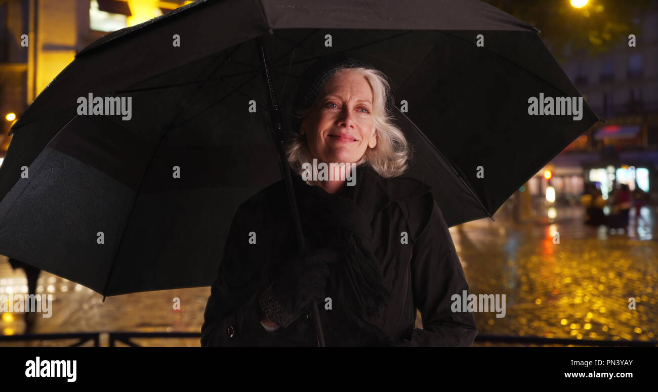 Happy senior woman in Paris smiling under umbrella on rainy night in ...