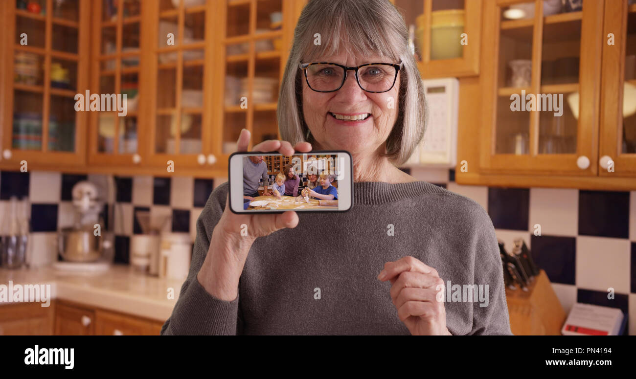 Happy white grandmother showing off video of family on mobile device in kitchen Stock Photo