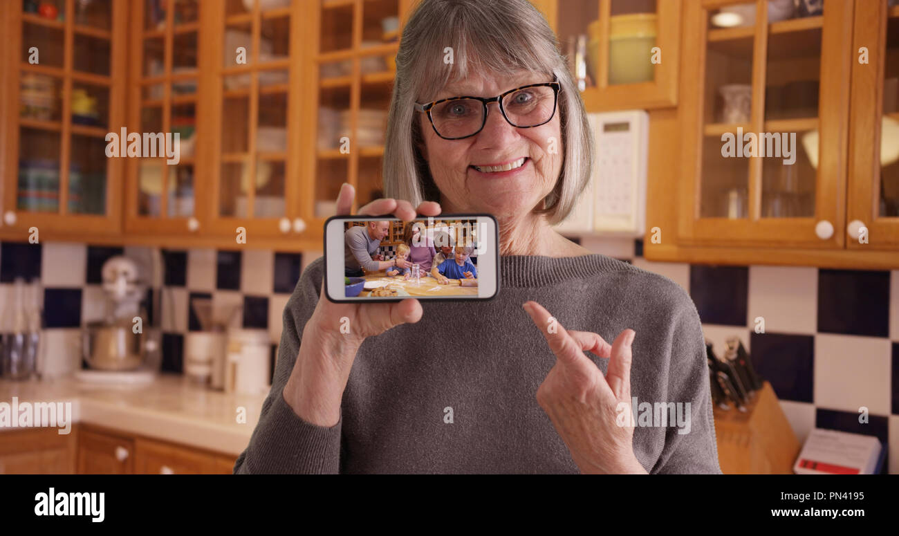 Happy white grandmother showing off video of family on mobile device in kitchen Stock Photo