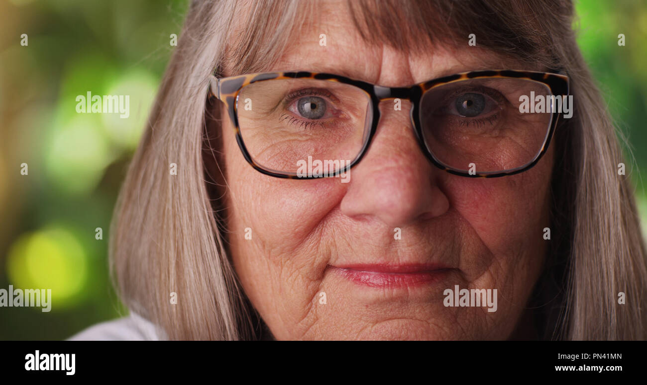 Tight shot of somber mature woman wearing eyeglasses outside in lush environment Stock Photo