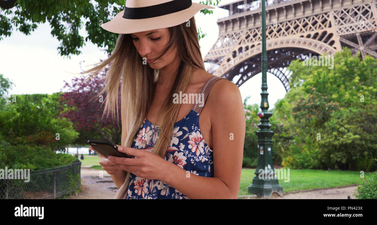 Millennial girl in romper and fedora texting on phone by Eiffel