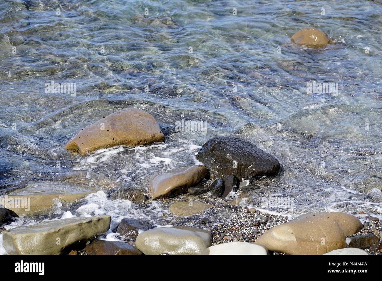 Large rounded rocks, small pebbles and shell grit at the edge of the sea water. Ripples in the shallow water. Stock Photo