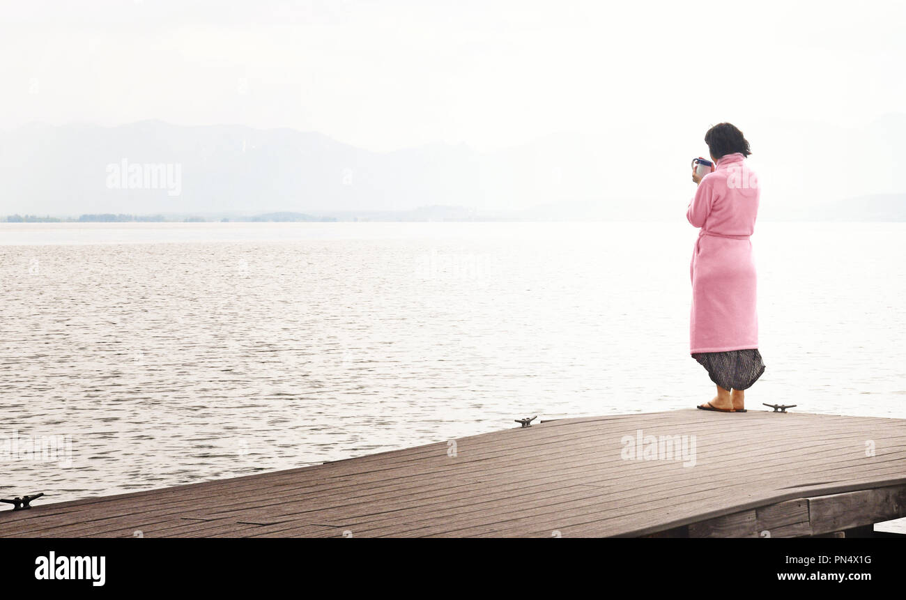 Middle aged caucasian woman standing on lakeside dock drinking cup of coffee on a stormy mountain morning Stock Photo