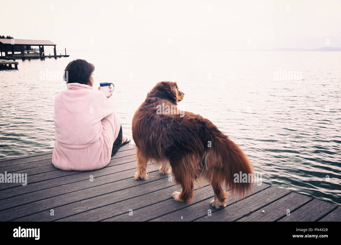 Middle aged caucasian woman and her dog sitting on lakeside dock drinking cup of coffee on a stormy mountain morning Stock Photo