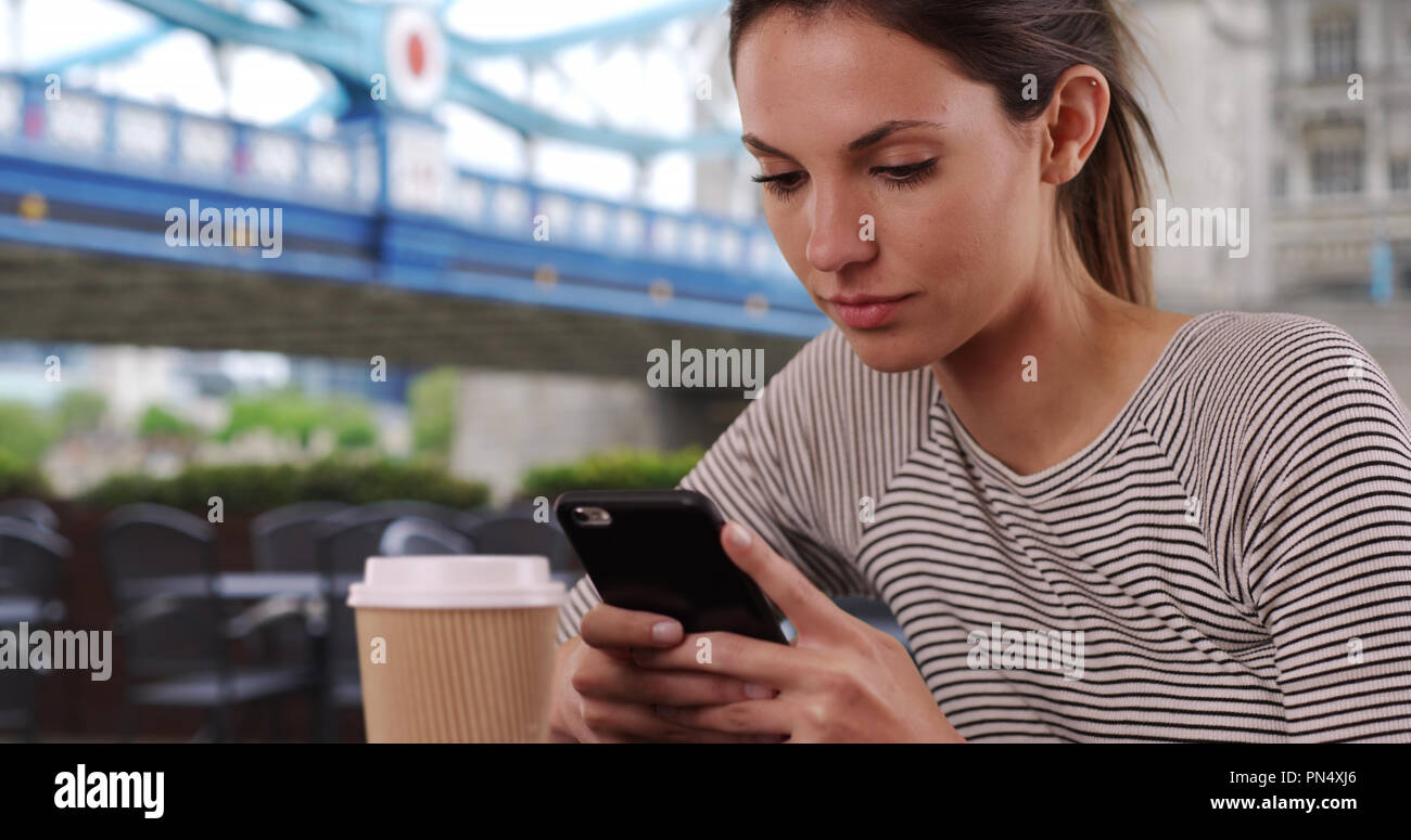 Young Caucasian lady with coffee cup messaging on phone seated by Tower Bridge Stock Photo