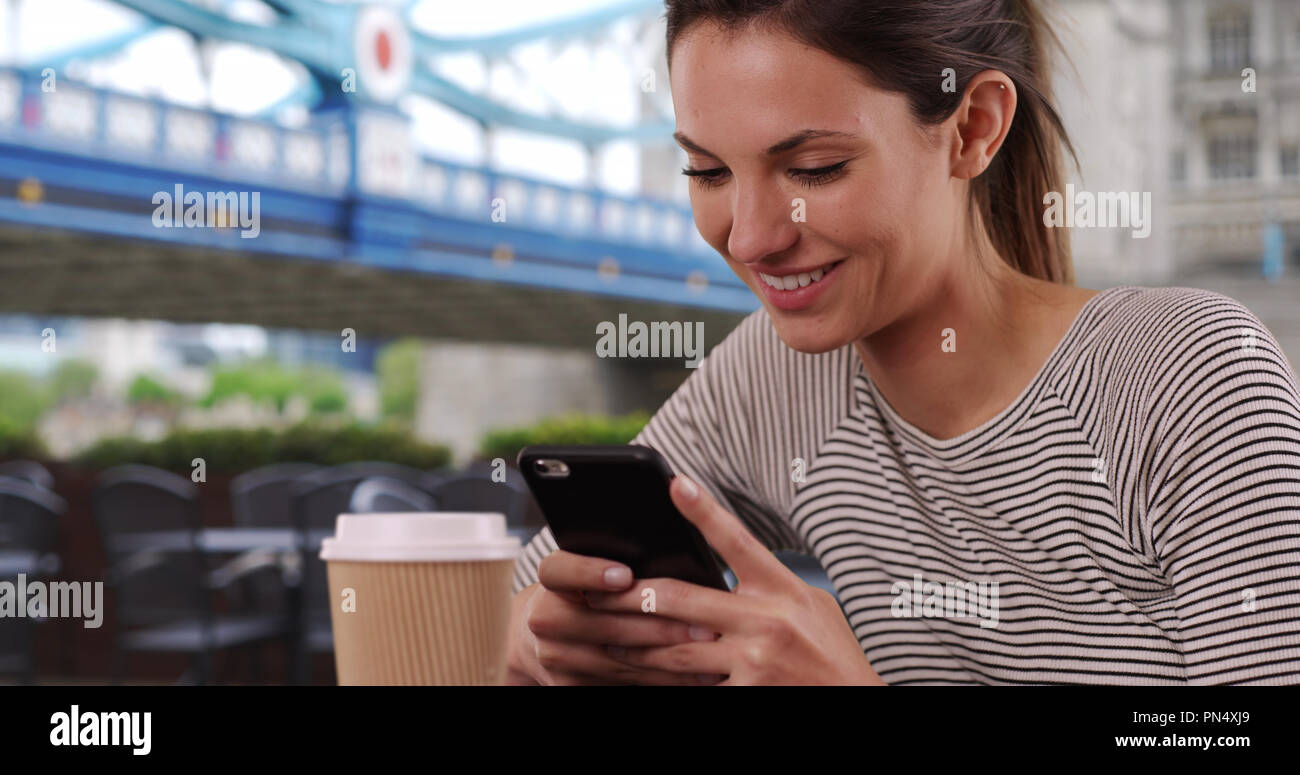 Young Caucasian lady with coffee cup messaging on phone seated by Tower Bridge Stock Photo