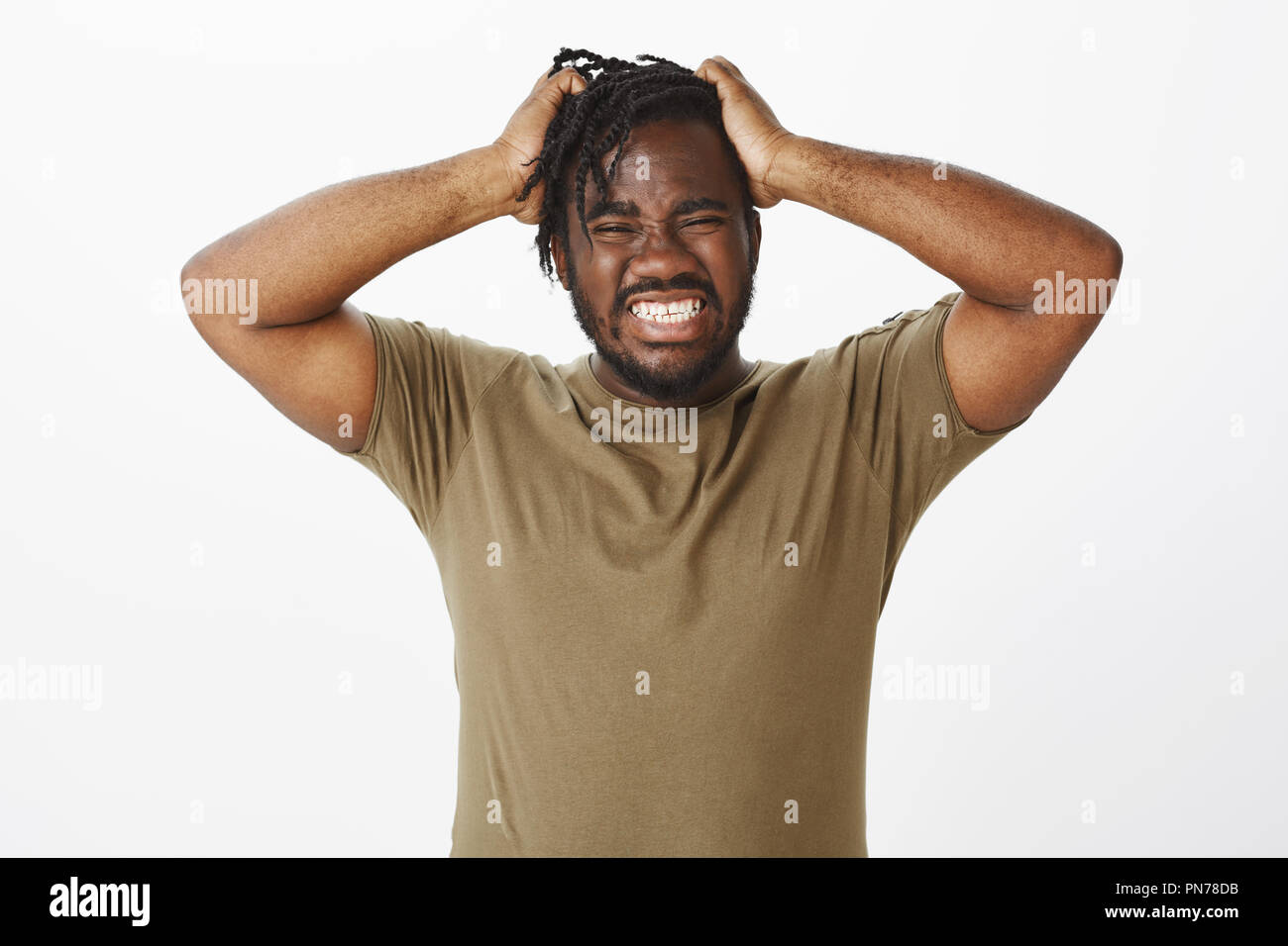 Guy cannot hold negative emotions inside. Depressed desperate african american man in olive t-shirt, holding hands on hair, grimacing with clenched teeth, feeling pain in soul or being angry Stock Photo