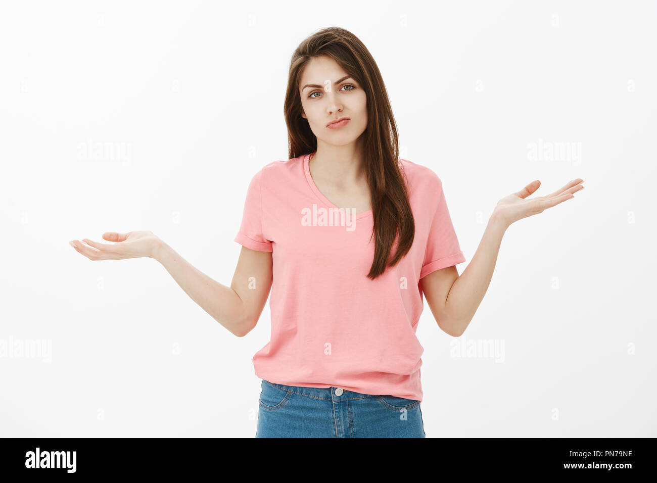 Indoor shot of displeased questioned cute brunette in pink t-shirt, spreading hands in clueless gesture, shrugging, being upset of losing, standing frustrated and gloomy over gray wall Stock Photo