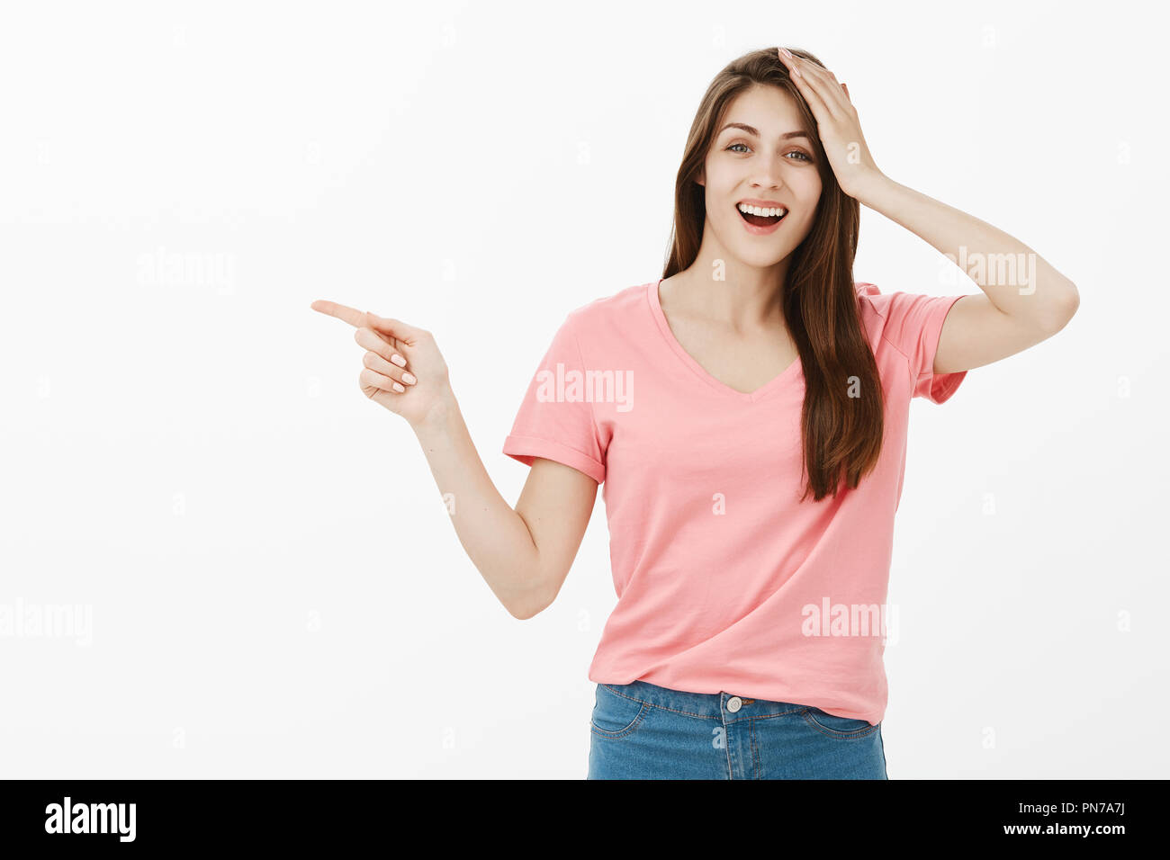 It totally slipped my mind. Portrait of confused happy good-looking woman in pink t-shirt pointing left with index finger while holding palm on head and smiling, being forgetful and asking question Stock Photo