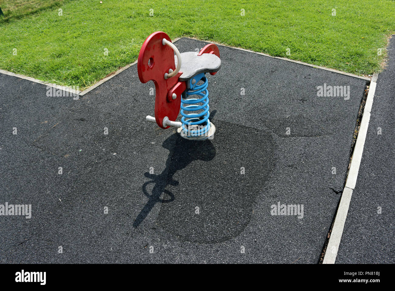 Red rocking bird piece of play equipment on a blue spring surrounded by a rubber crumb safety surface which has been repaired in several places and is Stock Photo