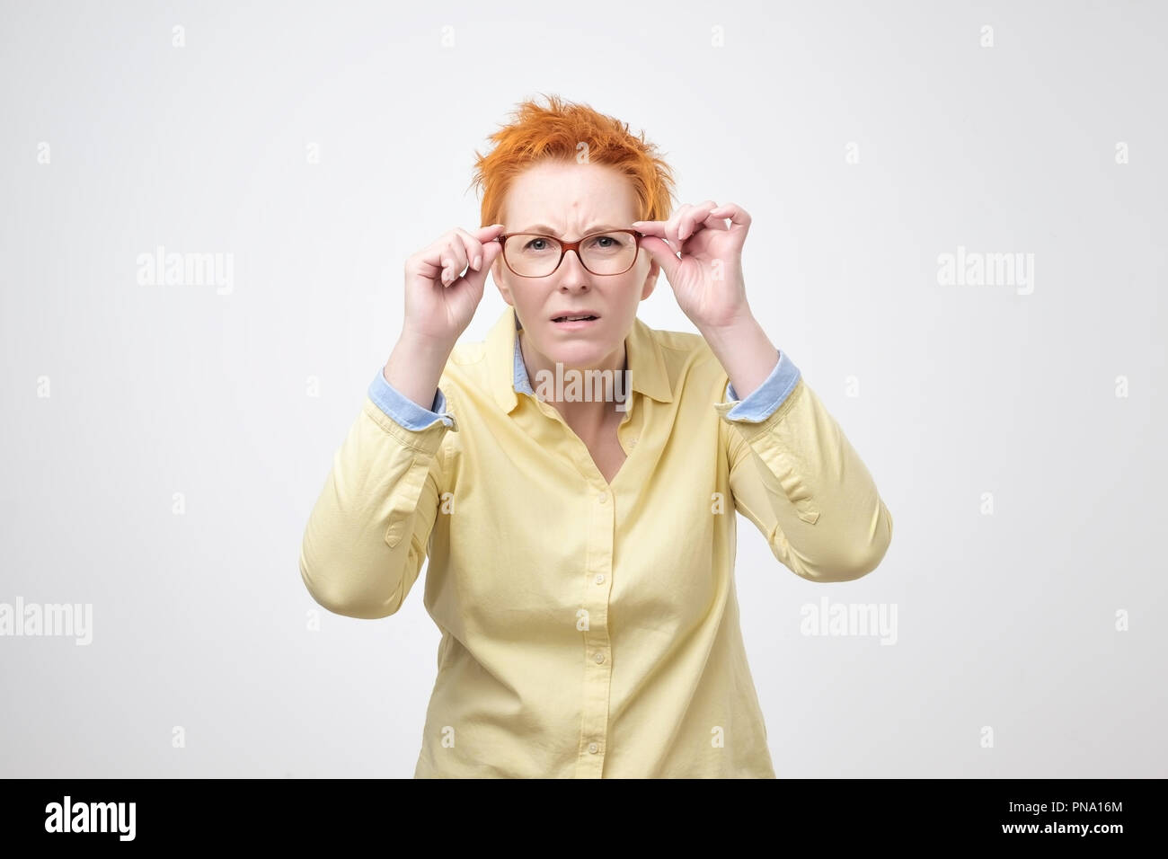 Attentive caucasian woman with short red hair looks scrupulously into distance, keeps hand on rim of spectacles. Try to see something far away, isolat Stock Photo