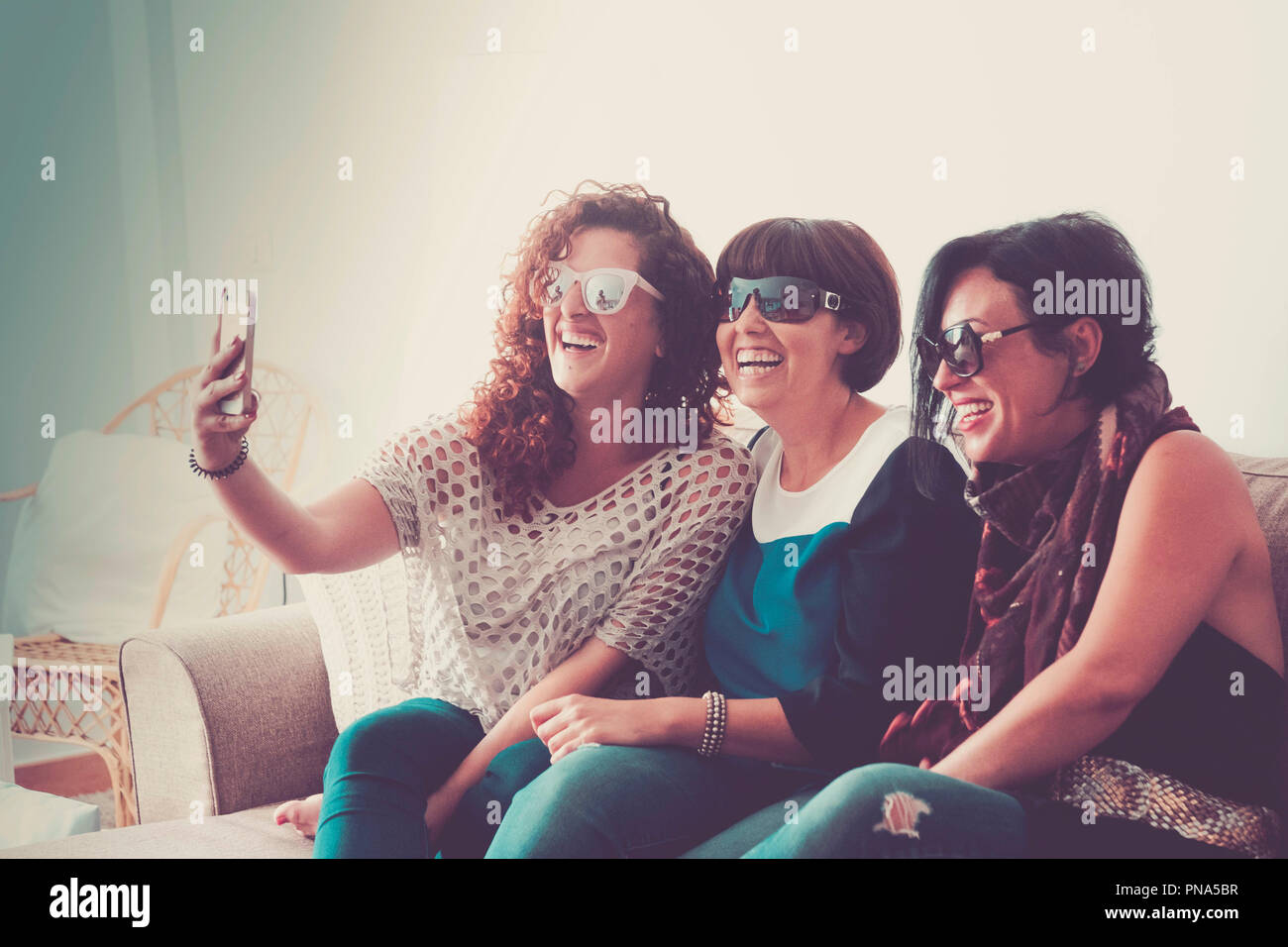 three caucasian young beautiful females doing a picture in selfie style with a mobile phone at home sit down on the sofa. friendship and connected wit Stock Photo