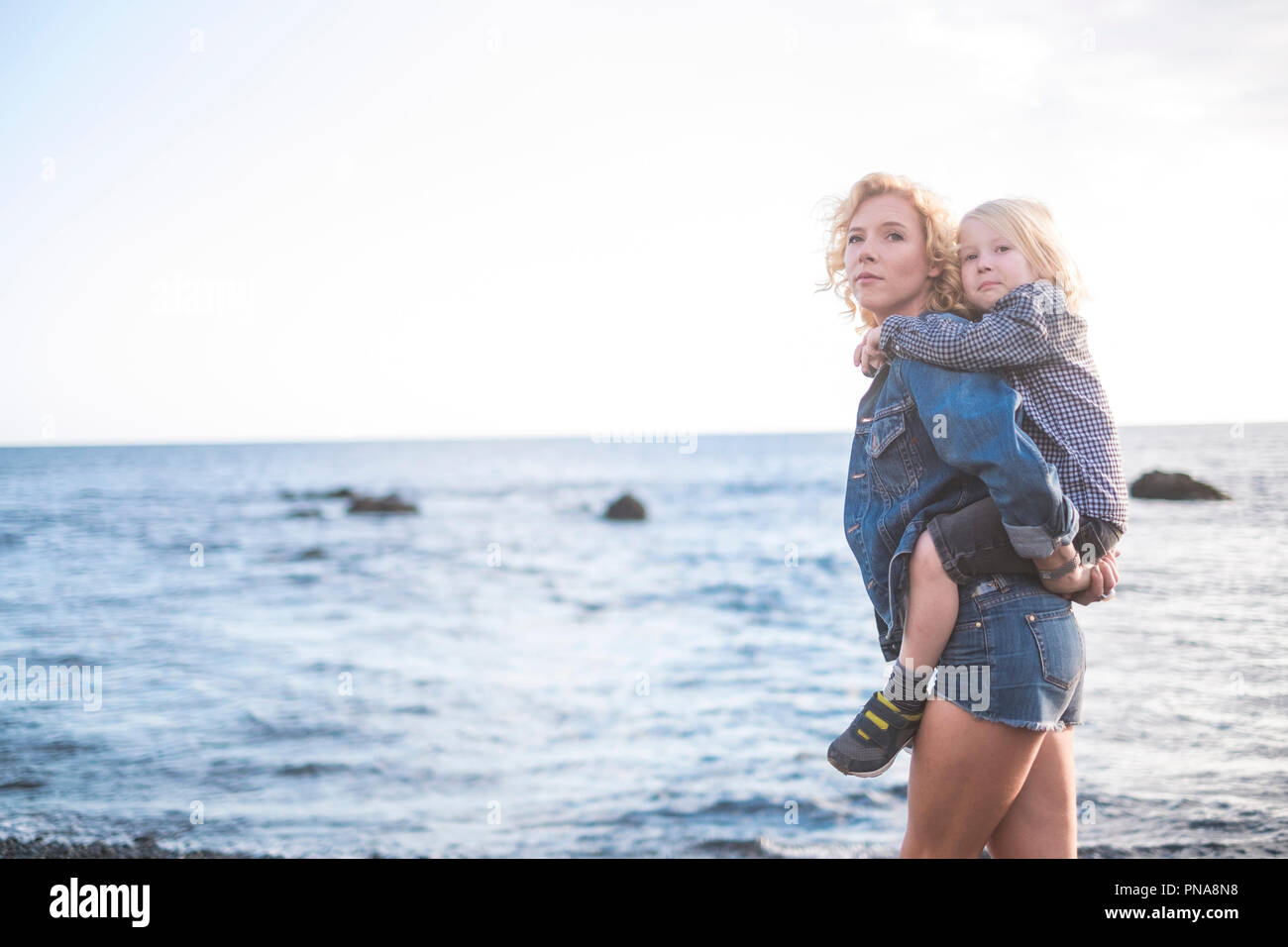beautiful young mother and little child blonde too walking at the beach in happiness. enjoying the outdoor activity and stay in love. the woman carry  Stock Photo