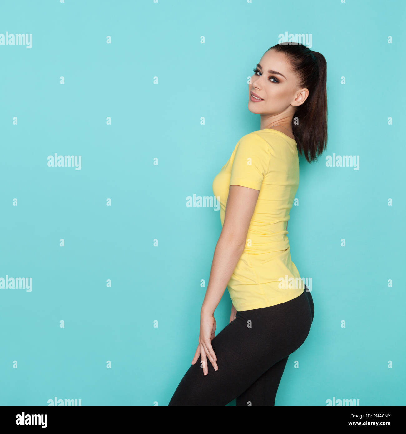 Download Beautiful Young Woman In Yellow Shirt And Black Leggings Is Standing And Looking At Camera Side View Three Quarter Length Studio Shot On Turquoise B Stock Photo Alamy PSD Mockup Templates