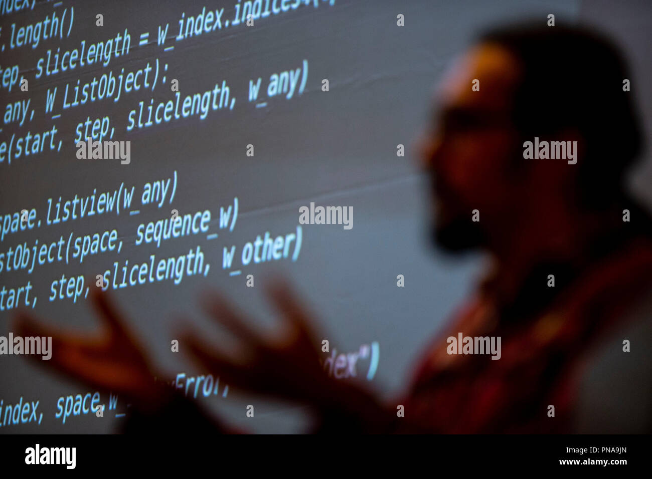 A man delivers a presentation in front of a screen of programmatic code. Stock Photo