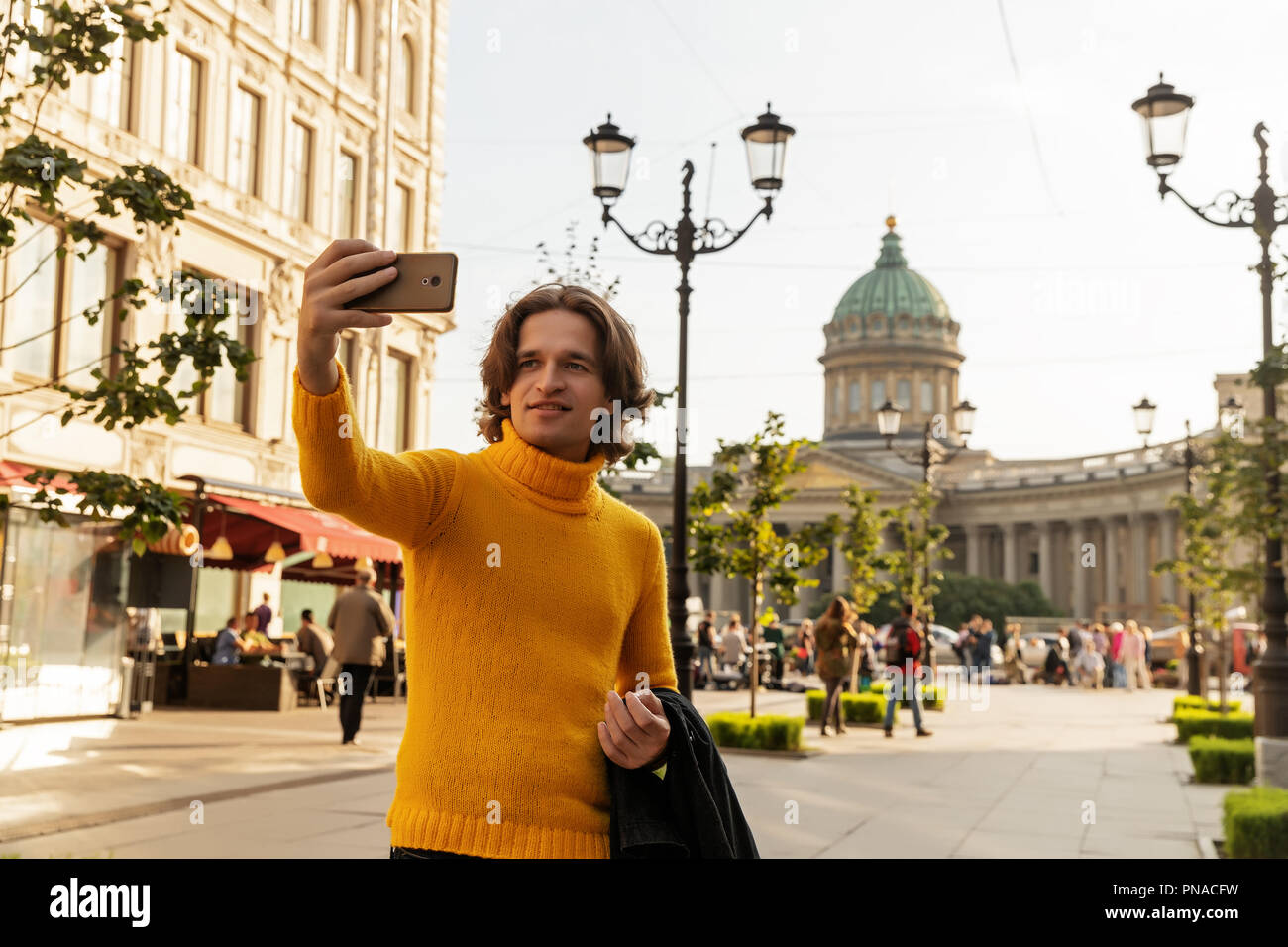 The young man waiting someone and does selfie, he dressed in a yellow sweater, a black raincoat or jacket, jeans, street and Kazanskiy cathedral Stock Photo