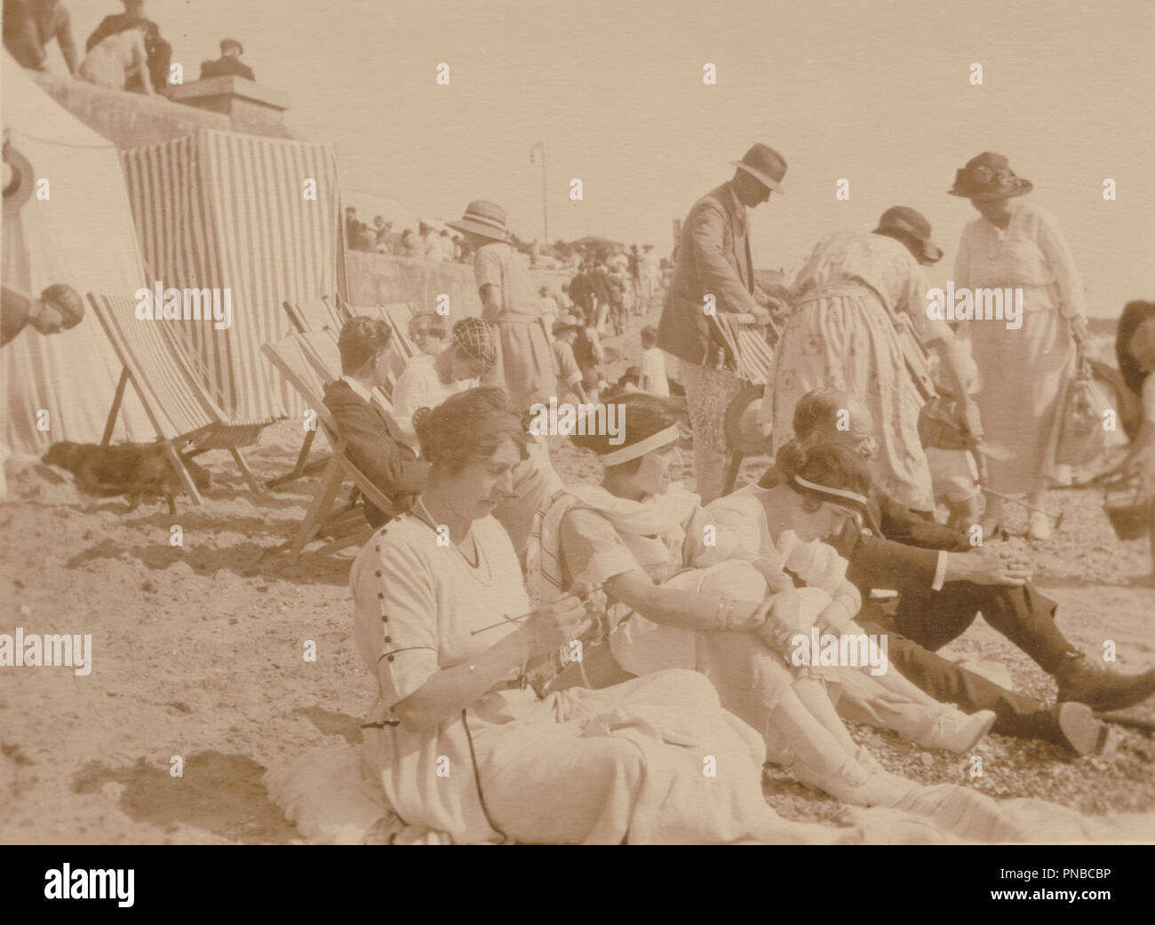 * From The Outfits Possibly 1920's Photograph of People at The Seaside Sitting On The Sandy Beach. One of The Ladies is Knitting. Stock Photo