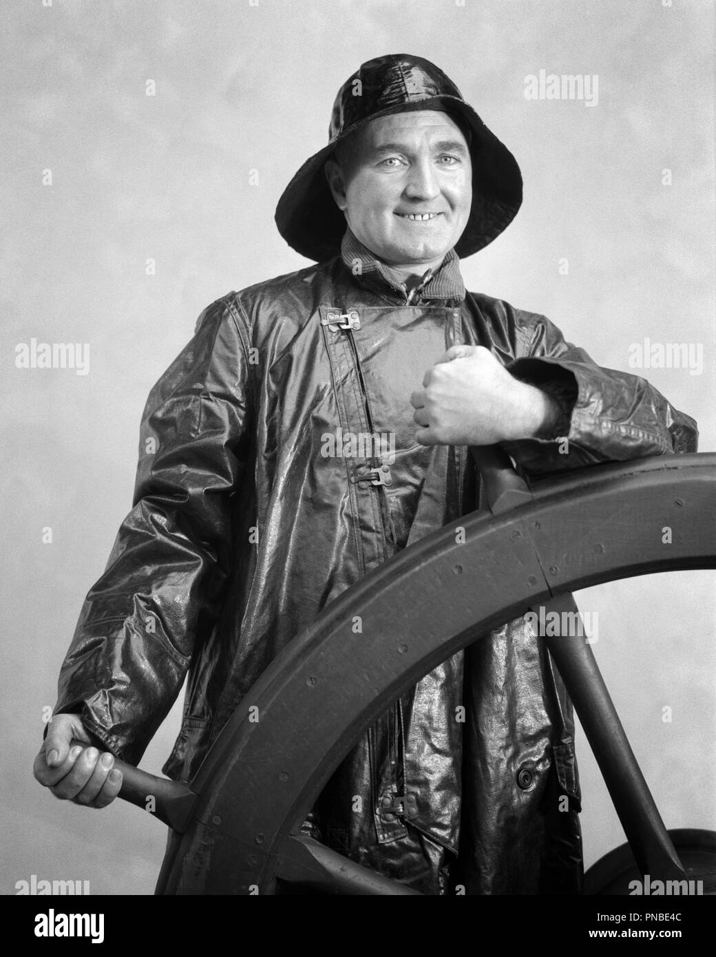1930s SMILING MAN SAILOR WEARING OILSKIN SOU’WESTER HAT AND RAIN COAT LOOKING AT CAMERA STANDING WATCH AT SHIP’S WHEEL - a2269 HAR001 HARS SAILOR JOBS HEALTHINESS HALF-LENGTH PERSONS DANGER MALES RISK CONFIDENCE EXPRESSIONS MIDDLE-AGED B&W MIDDLE-AGED MAN EYE CONTACT SKILL OCCUPATION HAPPINESS SKILLS CHEERFUL ADVENTURE HELM STRENGTH COURAGE AND EXPERIENCED EXCITEMENT KNOWLEDGE PROGRESS PRIDE AT GRIPPING OCCUPATIONS SMILES SPOKES JOYFUL SHIP'S OLD SALT ABLE CONFIDENT MID-ADULT MID-ADULT MAN OILSKIN BLACK AND WHITE CAUCASIAN ETHNICITY HAR001 OLD FASHIONED SEAMAN Stock Photo