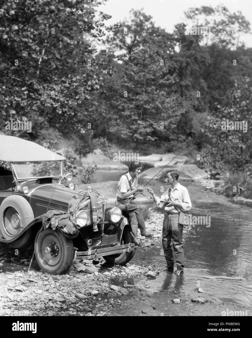 1920s COUPLE FLY FISHING MAN STANDING IN STREAM WOMAN SITTING  ON FENDER OF CAR   - a3527 HAR001 HARS TRANSPORTATION B&W HIGH ANGLE ADVENTURE LEISURE AUTOS RECREATION AUTOMOBILES ESCAPE STYLISH VEHICLES ANGLING FENDER MID-ADULT MID-ADULT MAN TOGETHERNESS YOUNG ADULT WOMAN BLACK AND WHITE CAUCASIAN ETHNICITY HAR001 OLD FASHIONED Stock Photo