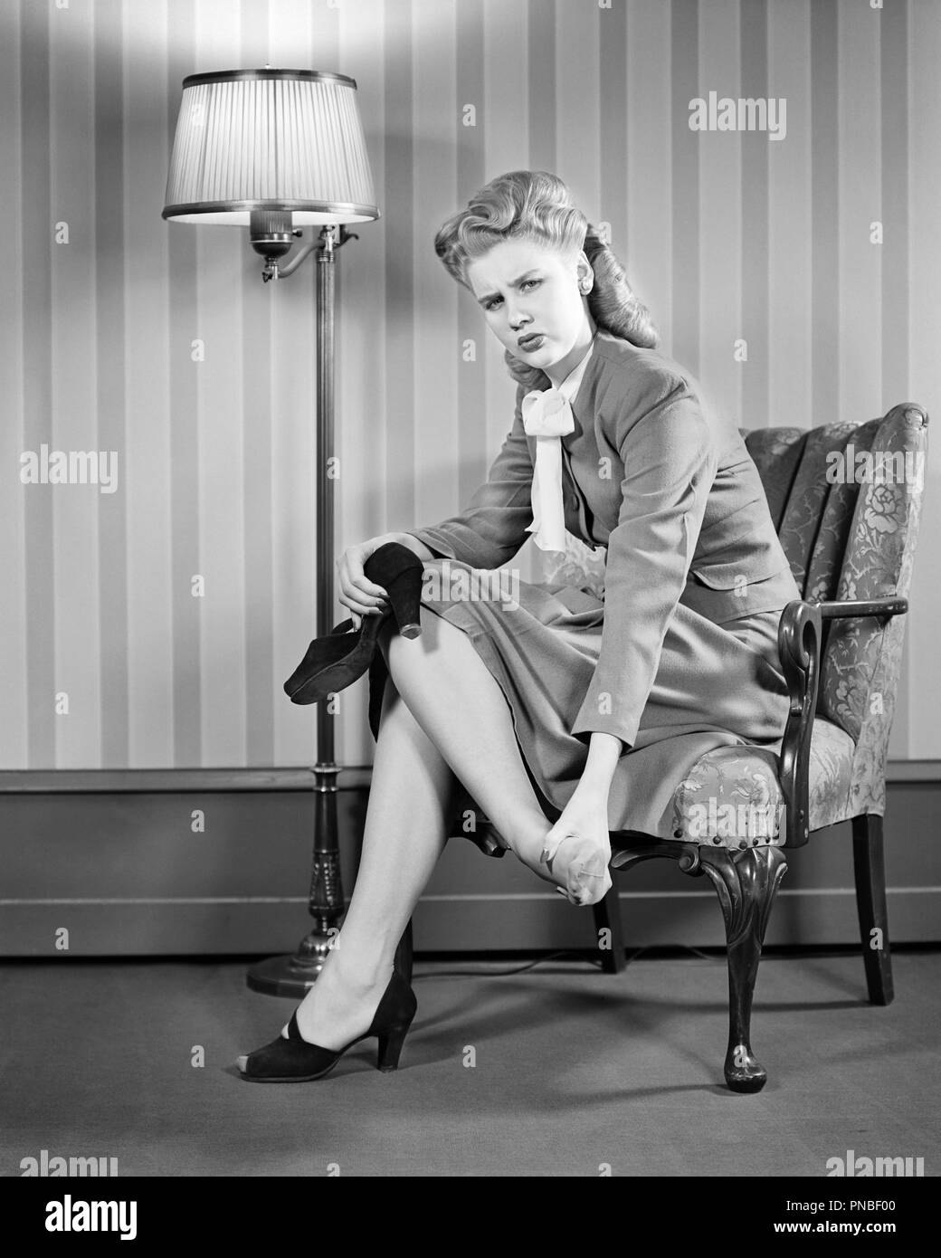 1940s FASHIONABLY DRESSED WOMAN BLOND VICTORY ROLLS HAIR STYLE LOOKING AT CAMERA SITTING ON LIVING ROOM CHAIR RUBBING SORE FOOT - a3869 HAR001 HARS PHYSICAL FITNESS PERSONS B&W SADNESS RUBBING EYE CONTACT TIGHT WELLNESS DISTRESSED STYLES LIVING ROOM HURT TOES UPDO FASHIONABLY VICTORY ROLLS STYLISH FASHIONS MASSAGING PAINFUL YOUNG ADULT WOMAN BLACK AND WHITE CAUCASIAN ETHNICITY HAIR STYLE HAR001 OLD FASHIONED Stock Photo