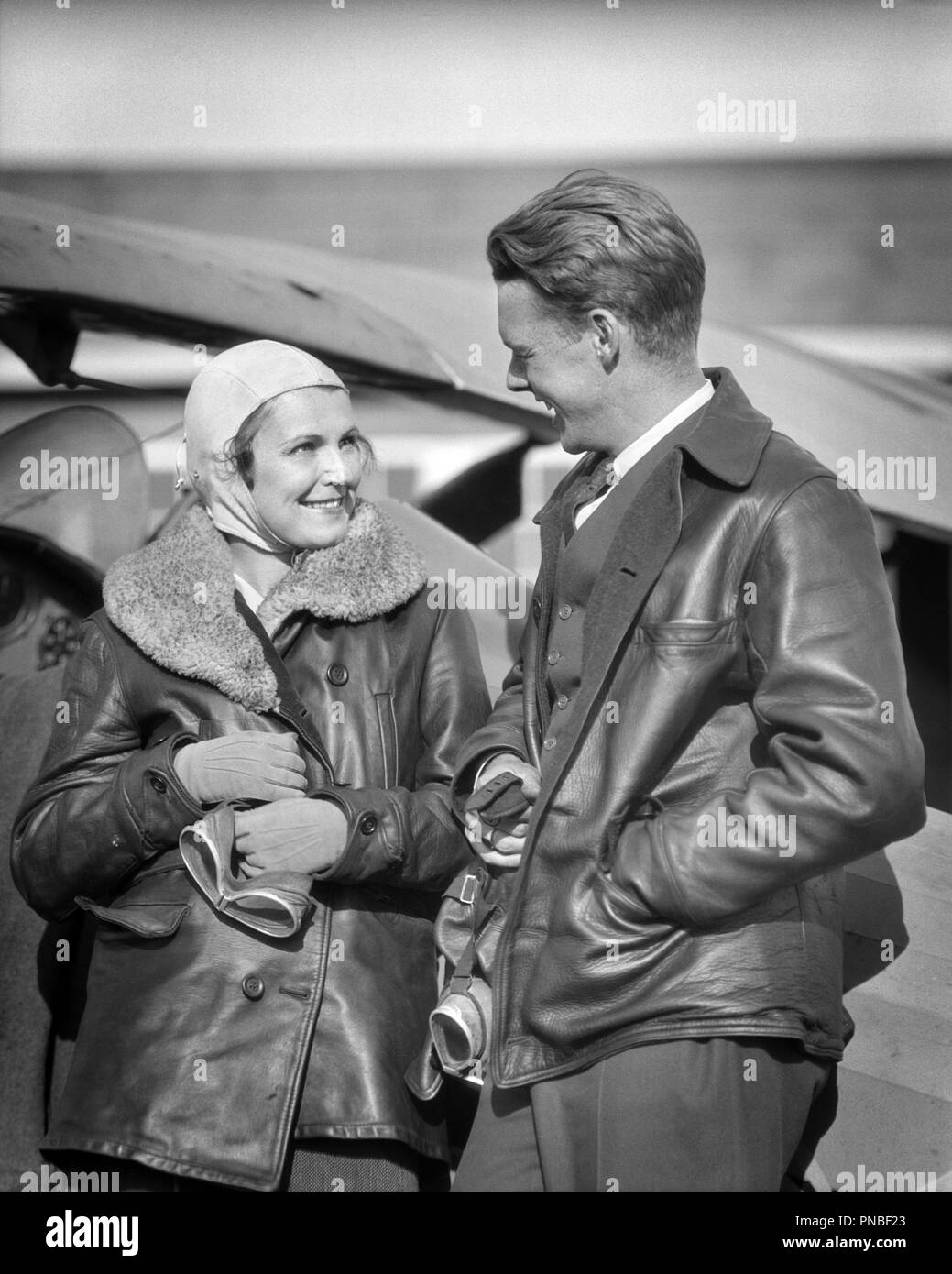 1930s COUPLE WEARING LEATHER JACKETS HOLDING GOGGLES LOOKING AT TALKING TO EACHOTHER STANDING BY AIRPLANE  - a4378 HAR001 HARS 1 AIRCRAFT FACIAL STYLE TEAMWORK PLEASED JOY LIFESTYLE PLANES FEMALES MARRIED SPOUSE HUSBANDS HEALTHINESS FRIENDSHIP HALF-LENGTH LADIES PERSONS GOGGLES MALES TRANSPORTATION EXPRESSIONS B&W PILOTS HAPPINESS CHEERFUL ADVENTURE AIRPLANES STYLES EXCITEMENT AT BY TO AVIATION OCCUPATIONS SMILES AVIATOR ESCAPE JOYFUL STYLISH AVIATRIX FASHIONS TOGETHERNESS WIVES BLACK AND WHITE CAUCASIAN ETHNICITY COPILOT EACH OTHER HAR001 JACKETS OLD FASHIONED Stock Photo