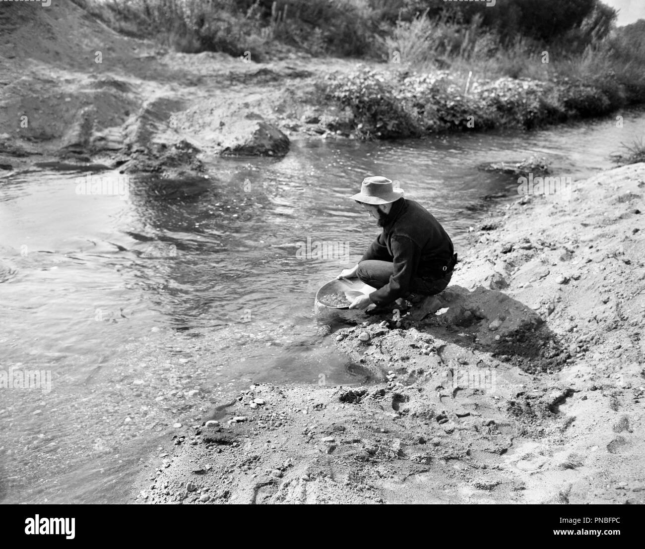 1900s MAN PROSPECTOR PANNING FOR GOLD IN STREAM - ap010254 CAM001 HARS ADVENTURE DISCOVERY PROSPECTOR LUCKY CAM001 MINING OCCUPATIONS GOLD RUSH PANNING SEARCHING GOLD MINE MID-ADULT MAN MOVIE STILL PROSPECTING BLACK AND WHITE CAUCASIAN ETHNICITY CREEK OLD FASHIONED OLD-FASHIONED Stock Photo
