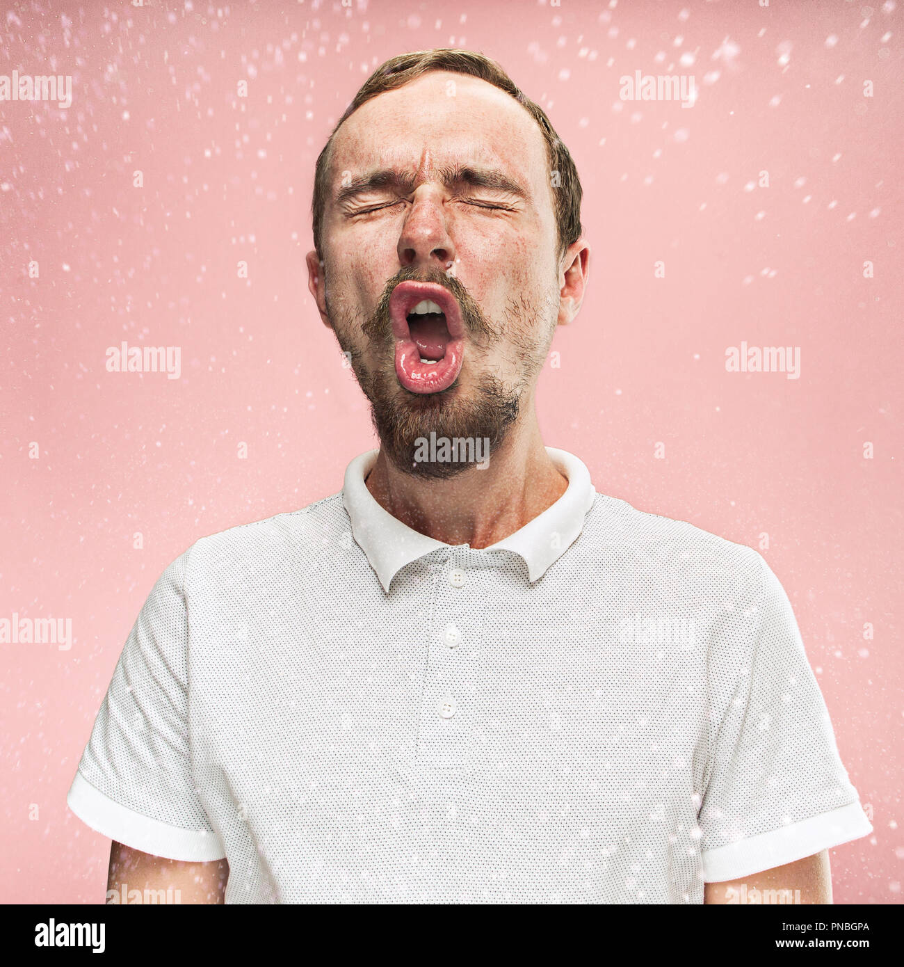 Young funny handsome man with beard and mustache sneezing with spray and small drops, studio portrait on pink background. Comic, caricature, humor. illness, infection, ache. Health concept Stock Photo